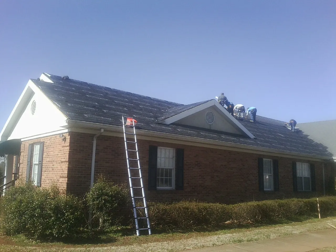 Crew tearing shingles off a brick ranch roof with a ladder set — Metal Roof Repair work by our crew in Clearbrook