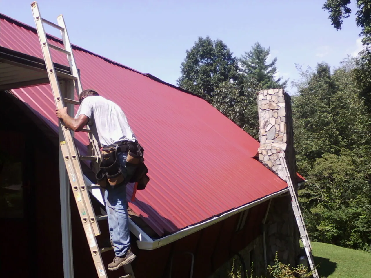 Roofer on a ladder beside a red metal roof and stone chimney — Soffit & Fascia Repair by Roofing Brothers in Clearbrook